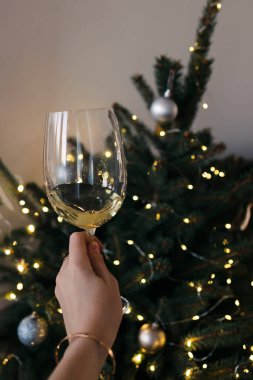 Woman's hand holding a glass of white or sparkling wine near the Christmas tree with lights