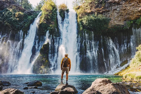 戴着帽子和黄色夹克的男人看着美丽的瀑布 从后面看去 — 图库照片