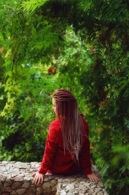 Yeşil çalıların yanındaki uzun saçlı kadın.