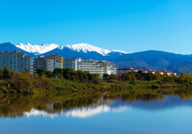 Önünde dalgalı bir göl, ortasında modern binalar olan karlı Kafkas dağlarının güzel gün ışığı manzarası. Sochi, Adler.