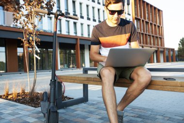 Alışveriş merkezinin yanında elektrikli scooterlı bankta oturan ve dizüstü bilgisayarla çalışan genç adam.