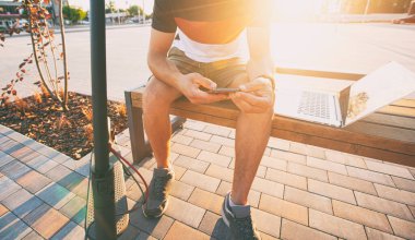 Alışveriş merkezinin yanında elektrikli scooterlı bankta oturan ve dizüstü bilgisayarla çalışan genç adam.