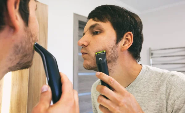 Young guy shaving with electric razor in the bathroom in front of the mirror