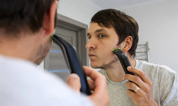 在镜子前的浴室里用电动剃须刀刮胡子的年轻人 — 图库照片
