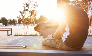 Alışveriş merkezinin yanında elektrikli scooterlı bankta oturan ve dizüstü bilgisayarla çalışan genç adam.