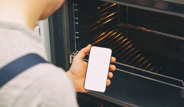 オーブンを修復するために来ている修理工の手に電話で白い空白の画面 スマートフォンの空のスペースでモックアップ 電話画面上のテキストの場所 — ストック写真