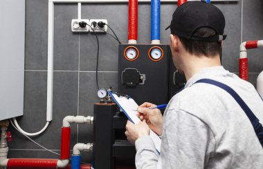 Technician servicing holding clipboard and inspecting heating system in boiler room