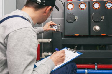Technician servicing holding clipboard and inspecting heating system in boiler room