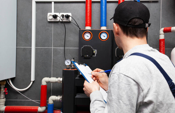 Technician servicing holding clipboard and inspecting heating system in boiler room