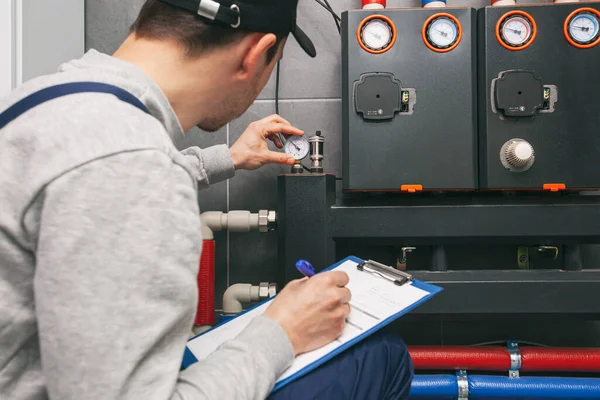 Technician servicing holding clipboard and inspecting heating system in boiler room