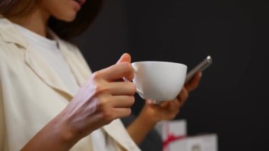 Genç bir kadın elinde beyaz bir fincan kahve tutuyor ve telefonuna yazıyor.