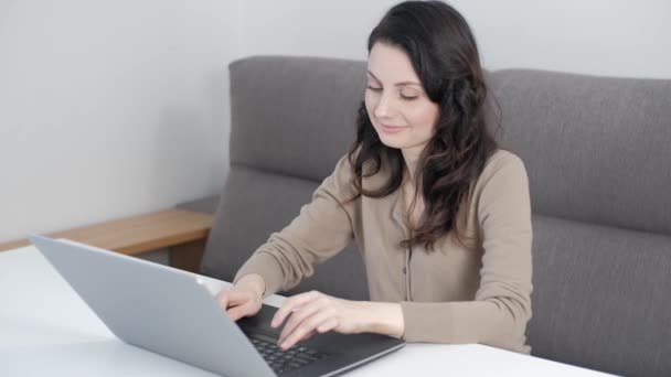 家庭でノートパソコンを使って働くフリーランスの女性 リビングルームの白いテーブルの後ろのソファに座っている間 ノートブックキーボードにテキストを入力するフリーランサー — ストック動画