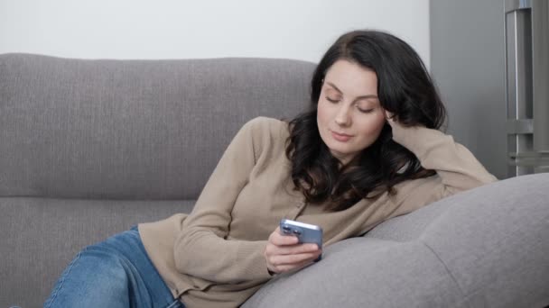 自宅のソファに横たわっている間 女性は携帯電話でニュースフィードをスクロールします スマートフォン上のソーシャルメディアアプリケーションを使用して30代の美しいブルネットモデルのストックビデオクリップ — ストック動画