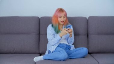 Mobil uygulamayla internetten saç boyalı alışveriş yapan harika bir kız. Evdeki koltukta otururken modern akıllı telefon cihazıyla sosyal medya uygulamalarını karıştıran renkli saçlı genç bir kadın.