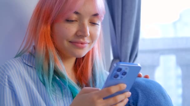 染め髪の若い女性は 携帯電話でソーシャルメディアニュースフィードを参照してください 現代のスマートフォンのモバイルアプリを使って髪の色をした可愛い女の子 4Kストックビデオのかなり白い女性とともに虹の髪型 — ストック動画