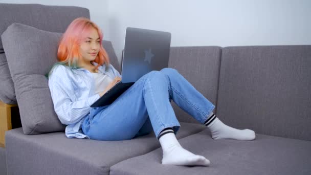 在手提电脑上输入染发的小女孩 自由职业女性的股票视频 她的头发是彩色的 在家里被锁起来 躺在她的公寓沙发上 免费提供4K胶卷 — 图库视频影像