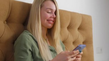Güzel, sarışın bir kadın cep telefonu kurye uygulamasında mutlu bir gülümsemeyle mesaj yazıyor. Çevrimiçi iletişim için modern akıllı telefonu kullanan beyaz bir kadının video klibi.