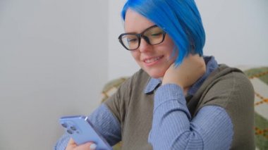 Cep telefonu kullanan mutlu bir milenyum kadını. Neşeli beyaz kadın, boyalı mavi saçlarıyla interneti tarıyor ve sosyal medya haberleri modern akıllı telefon cihazıyla besleniyor. Çevrimiçi iletişim kuran gülümseyen kişi