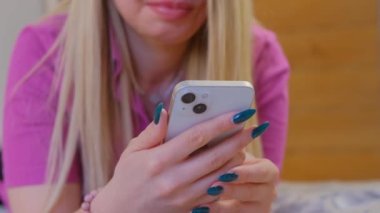 Sarışın kadın çift kameralı yeni akıllı telefon kullanıyor. Orkide rengi tişörtlü genç bir kadın yatak odasında yatarken modern beyaz cep telefonuyla internette geziniyor, 4k ultra HD ile çekiliyor.