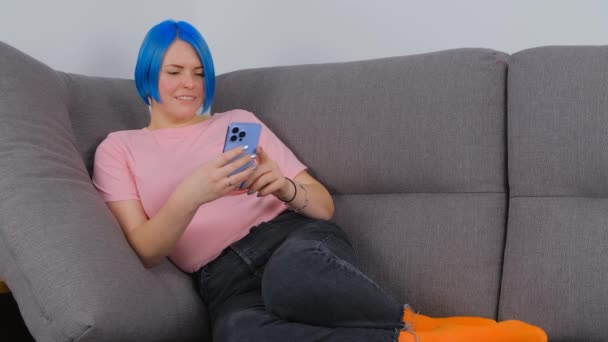 ソファの上で携帯電話を使って幸せな白い女性 モダンなスマートフォンで青髪のブラウジングインターネットを染めたリラックスした若い女性 カラーヘアコミュニケーションをオンラインで行う個人 — ストック動画