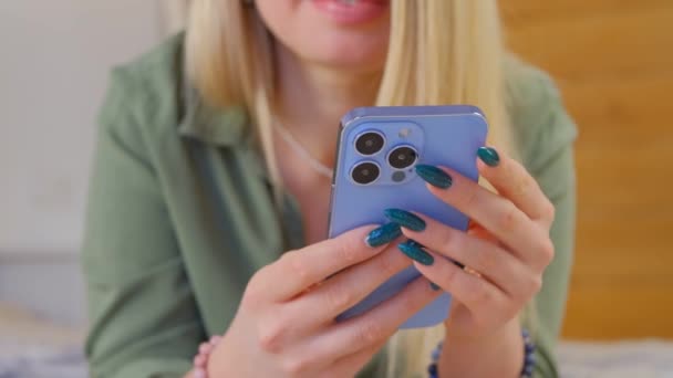 身穿橄榄绿衬衫的金发年轻女子躺在床上 用现代浅蓝色手机和三重摄像头 白人女性在智能手机上用4K视频浏览互联网 — 图库视频影像