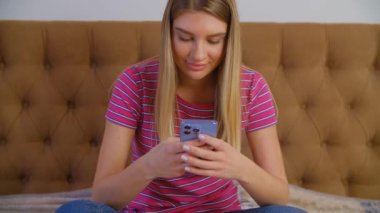 Neşeli beyaz kadın, yatak odasında kilit altında otururken mobil kurye uygulaması ile modern akıllı telefondan mesaj yazıyor. Güzel beyaz kadın iletişim için modern cep telefonunu kullanıyor.