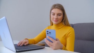 Güzel beyaz kadın tecrit sırasında evden bağımsız çalışıyor. Genç, sarışın bir kadın dizüstü bilgisayarın önünde neşeli bir gülümsemeyle modern cep telefonu uygulamasına göz atıyor. Serbest çalışan biri.