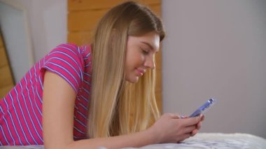 Güzel sarışın kadın yatak odasında kilit altında yatarken cep telefonundan mesaj yazıyor. Sosyal medya uygulamasında modern akıllı telefonu kullanarak 4k video kullanan mutlu beyaz kadın.