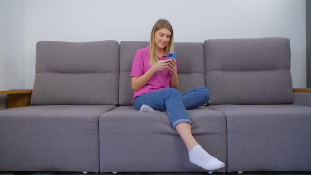 美しい若い女性は 通信のための携帯電話アプリを使用して ハッピーホワイト女性とともに長いですブロンド髪タイピングメッセージでメッセンジャーアプリケーションで現代的なスマートフォン上で座っている間ソファに家 — ストック動画