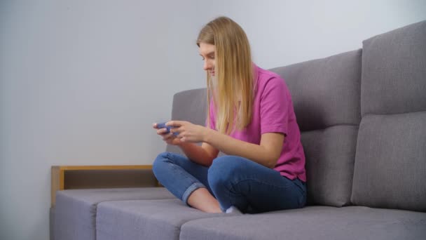 ゲーマーの女性は スマートフォンでビデオゲームをプレイ 蘭ポロシャツの興奮した白い女性は自宅でリビングルームでソファに座っている間 現代の携帯電話デバイス上で現代的なゲームをプレイ — ストック動画