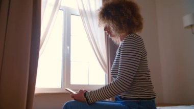 Yatak odasında cep telefonu kullanan kıvırcık saçlı genç bir kadın. Kıvırcık beyaz kadın yatakta otururken sosyal medya uygulamasında modern akıllı telefonla mesaj yazıyor.