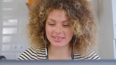Dizüstü bilgisayarında kulaklığı ve web kamerasıyla video görüşmesi yapan mutlu kıvırcık kadın. İnternet bağlantısı ve kulaklıkla iletişim kuran mutlu beyaz kadın. Serbest çalışan kişi müşteriyle çevrimiçi konuşuyor