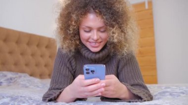 Güzel beyaz kadın evde yatağında uzanırken modern cep telefonuyla mesaj yazıyor. Tecrit sırasında iletişim için akıllı telefon ve hızlı internet kullanan çekici genç bir kadın.