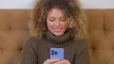 Mutlu beyaz kadın tecrit sırasında sosyal medya uygulamasını çevrimiçi iletişim için kullanıyor. Modern akıllı telefon cihazına mesaj yazan neşeli, genç bir kadın. Elinde cep telefonu olan güzel kıvırcık insan.