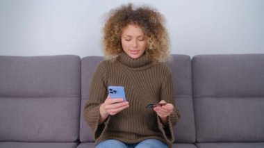 Mutlu beyaz kadın internetten kredi kartı ve modern cep telefonuyla alışveriş yapıyor. Güzel kıvırcık saçlı kadın banka kartı ve akıllı telefonuyla ödeme yapıyor. Neşeli insan telefonla para transferi yapıyor.