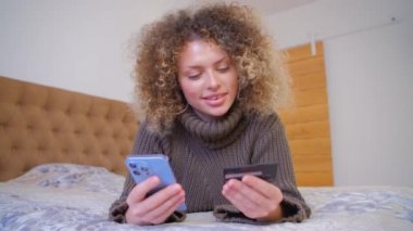 İnternette kredi kartıyla ödeme yapan mutlu bir alıcı. Kıvırcık beyaz kadın internet mağazasında ödemesini modern akıllı telefondan yapıyor. Neşeli müşteri çevrimiçi cep telefonu ve internet bağlantısı ile alışveriş yapıyor