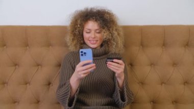 Akıllı telefon uygulaması ile kredi kartıyla ödeme yapan mutlu alıcı kadın. Neşeli beyaz kadın evde online alışveriş için modern mavi cep telefonunu kullanıyor. İnternet mağazasından kıvırcık saçlı güzel biri satın alıyor.