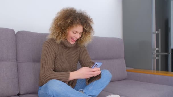 美しい幸せな女性が携帯電話アプリにメッセージを入力します ソファに座っている間明るい笑顔で現代のスマートフォン上のソーシャルメディアアプリケーションで巻き毛書きコメントを持つ若い大人の女性 — ストック動画