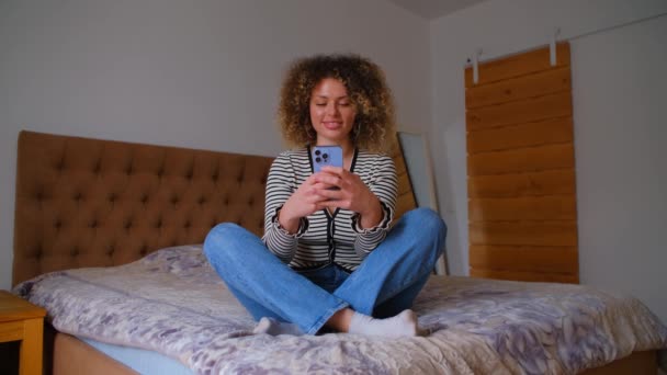 快乐的年轻女人在智能手机上输入消息 漂亮的卷曲女性在锁门时坐在卧室里 用现代手机进行交流 快乐的人在网上发短信 — 图库视频影像