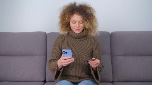 快乐的白人妇女用信用卡和现代手机在网上购物 漂亮的卷曲白色女性用银行卡和智能手机付款 快乐的人通过电话汇款 — 图库视频影像