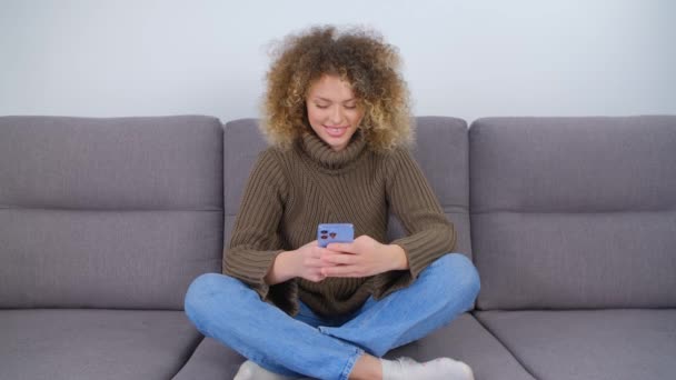 現代の携帯電話にメッセージを入力して幸せな白い女性 携帯電話を使って巻き毛のきれいな白人女性 ソファに座っている間に新しいスマートフォンでオンラインで通信する陽気な人 — ストック動画