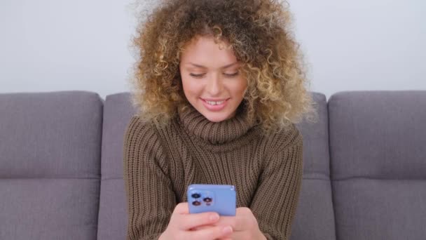 漂亮的年轻白人妇女在社交媒体应用程序上通过手机在线输入消息 快乐女性与卷发使用现代蓝色智能手机与三重相机沟通锁定 — 图库视频影像