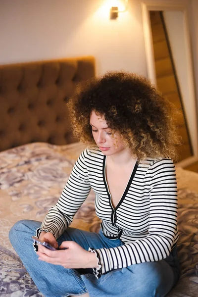 有卷发的年轻女人在卧室里用手机 坐在床上用现代智能手机在社交媒体应用程序上输入卷曲的白色女性信息 — 图库照片
