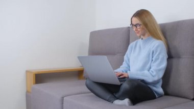 Dizüstü bilgisayarda yazan neşeli genç bir kadın. Genç sarışın kadın evde serbest çalışıyor. Gülümseyerek. 4k video 'da apartmandaki koltukta otururken bilgisayar üzerinde çalışan girişimci