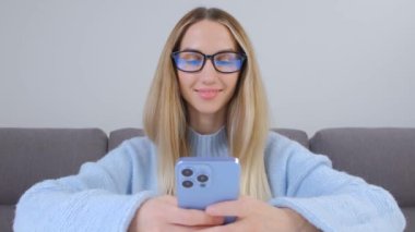 Cep telefonu kullanan mutlu genç bir kadın. Gözlüklü güzel sarışın kadın modern mavi akıllı telefonunda üçlü kamerayla mesaj yazıyor. Neşeli genç yetişkin insan cep telefonuyla iletişim kuruyor.