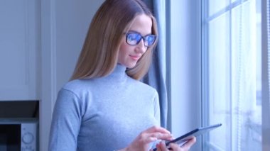 Evdeki pencerenin yanında tablet kullanan genç bir kadın. Gözlüklü güzel sarışın kadın internet haberlerini 4K video 'da modern mobil cihazla izliyor. Çevrimiçi iletişim ve eğlence kavramı