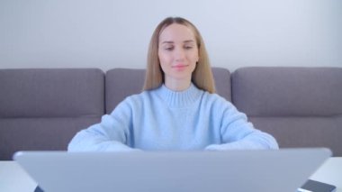 Kilitleme sırasında evde bilgisayar başında çalışan mutlu genç bir kadın. Uzun sarı saçlı neşeli beyaz kadın uzaktan gülümseyerek çalışıyor. Laptop klavyesine yazı yazan odaklanmış serbest yazar