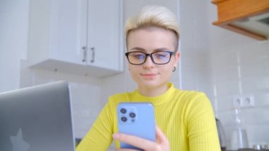 Çevrimiçi ağ kavramı. Tom Boy saç kesimli genç bir kadın müşterilerle iletişim kurmak için modern mavi cep telefonu ve dizüstü bilgisayar kullanıyor. Kilitleme sırasında evde çalışan serbest çalışan kişi 4K görüntülerinde