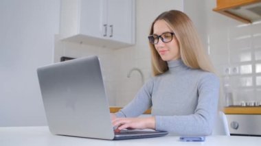 Güzel beyaz kadın evdeki dizüstü bilgisayarda çalışıyor. 4K video 'da not defterine uzun sarı saçlı ve yazı yazan çekici genç bir kadın. Serbest çalışan kişi uzak iş yapıyor.
