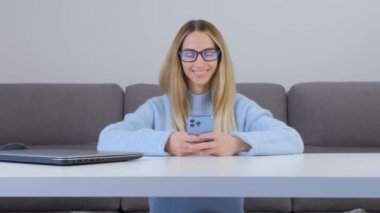 Cep telefonundaki kurye uygulamasında mesaj yazan mutlu sarışın kadın. Gözlüklü, genç ve güzel bir kadın. Evdeki iletişim için üç kameralı modern mavi akıllı telefon cihazı kullanıyor.